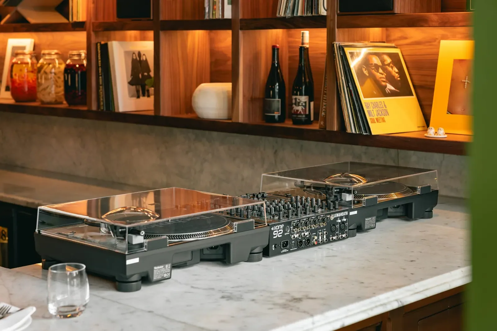 Turntables and a mixer at the end of the marble counter, sleeves and bottles on the walnut shelf behind.